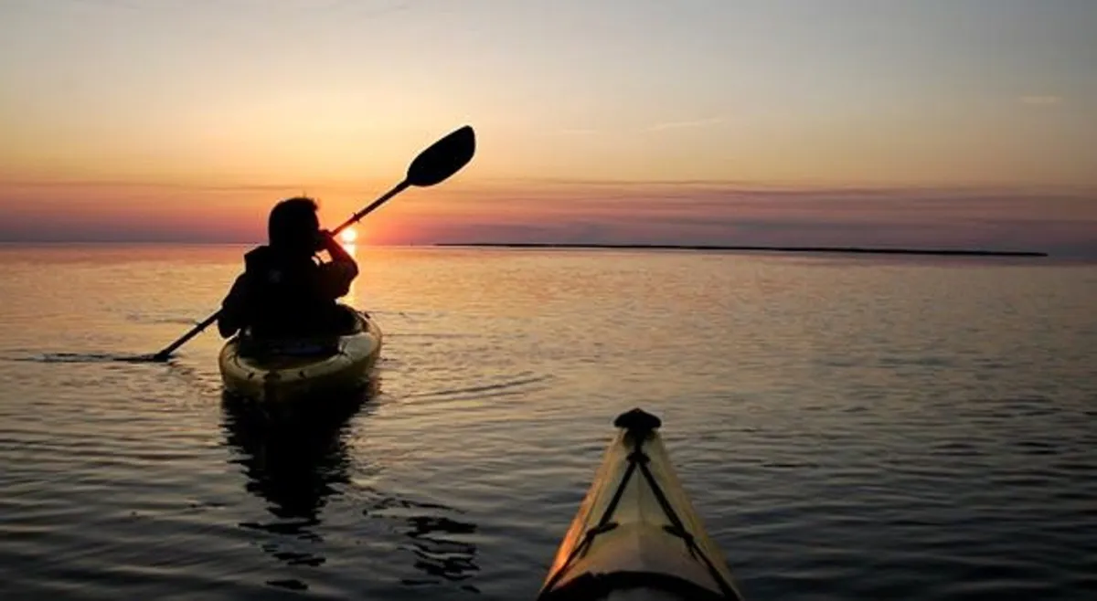 A picturesque scene of kayakers gliding across calm water at sunset, with the sky painted in warm hues and the surface of the sound reflecting the fading light. The mood is peaceful and intimate, ideal for shared moments.