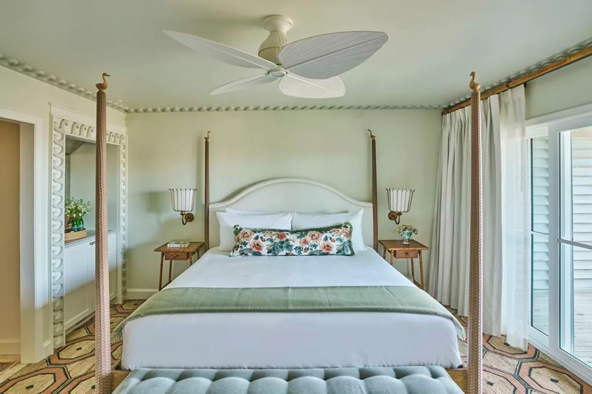A tranquil guestroom featuring a four-poster king bed adorned with decorative pillows, complemented by a soft ceiling fan and an elegant mirror. The room is bathed in gentle natural light, evoking a sense of calm and restful privacy.
