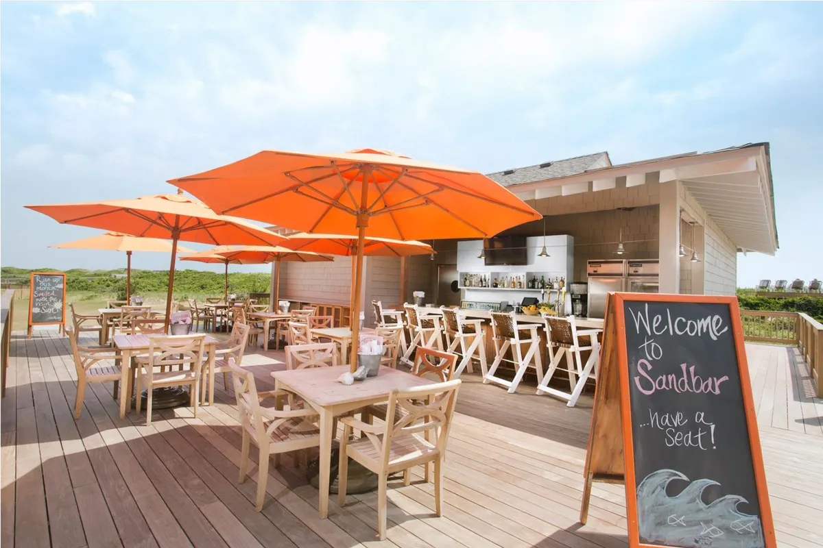 This exterior scene features vibrant orange umbrellas shading wooden tables in a relaxed barbecue and lounge area. The welcoming sign, open seating, and casual setup evoke a cheerful retreat atmosphere, inviting guests to unwind, socialize, or enjoy outdoor dining. The space is surrounded by greenery and bathed in natural light, suggesting a peaceful setting ideal for reflection or wellness activities.