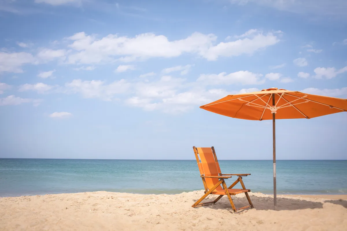 This image captures a tranquil beach scene, featuring an orange beach chair and umbrella set on soft sand with the blue ocean stretching into the distance. The relaxed setup invites thoughts of sunbathing, reading, or simply soaking in the calm seaside atmosphere.