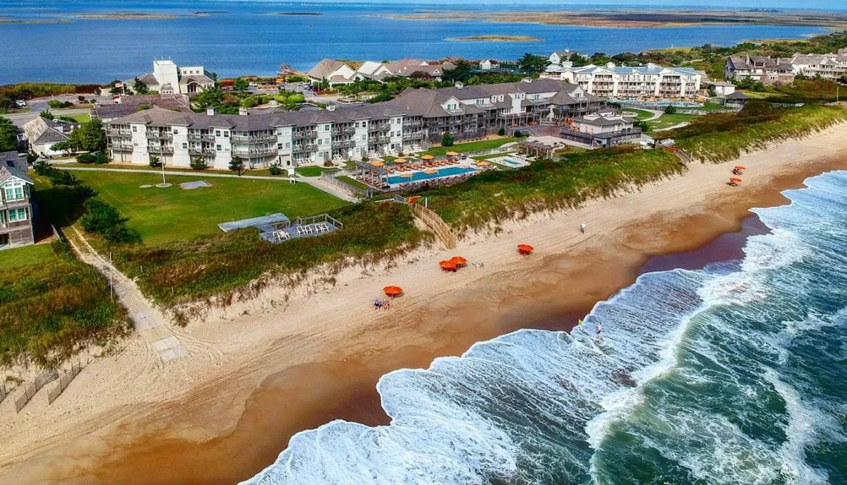 An inviting exterior shot of the resort's beachfront with a pristine swimming pool, sun loungers under umbrellas, and gentle ocean waves in the background. The image captures both the pool and beachfront leisure activities.