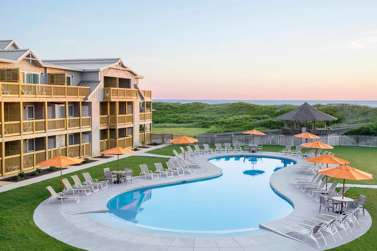 This exterior image showcases a sparkling swimming pool bordered by orange umbrellas, lush green grass, and visible hotel balconies. The ambiance is relaxed and casual, suggesting a welcoming space just steps from the accommodations. The gazebo and the open sky hint at both sunrise and sunset enjoyment, while the proximity of balconies suggests potential direct access from private outdoor spaces.