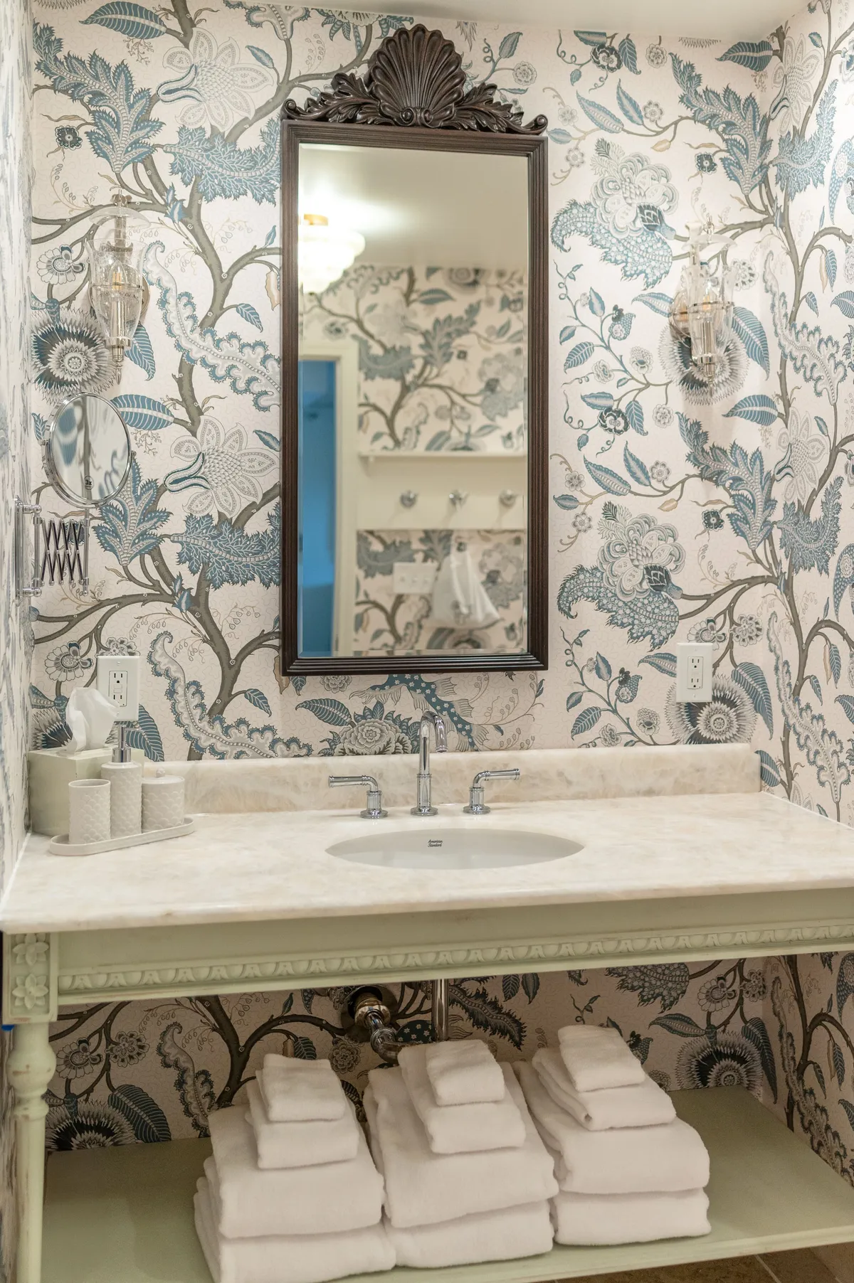 The image showcases a refined interior bathroom scene, featuring an ornate mirror, elegant floral wallpaper, a gleaming marble countertop, and freshly stacked towels. The space radiates tranquility, inviting guests to indulge in moments of self-care, with soft natural light enhancing the serene atmosphere.