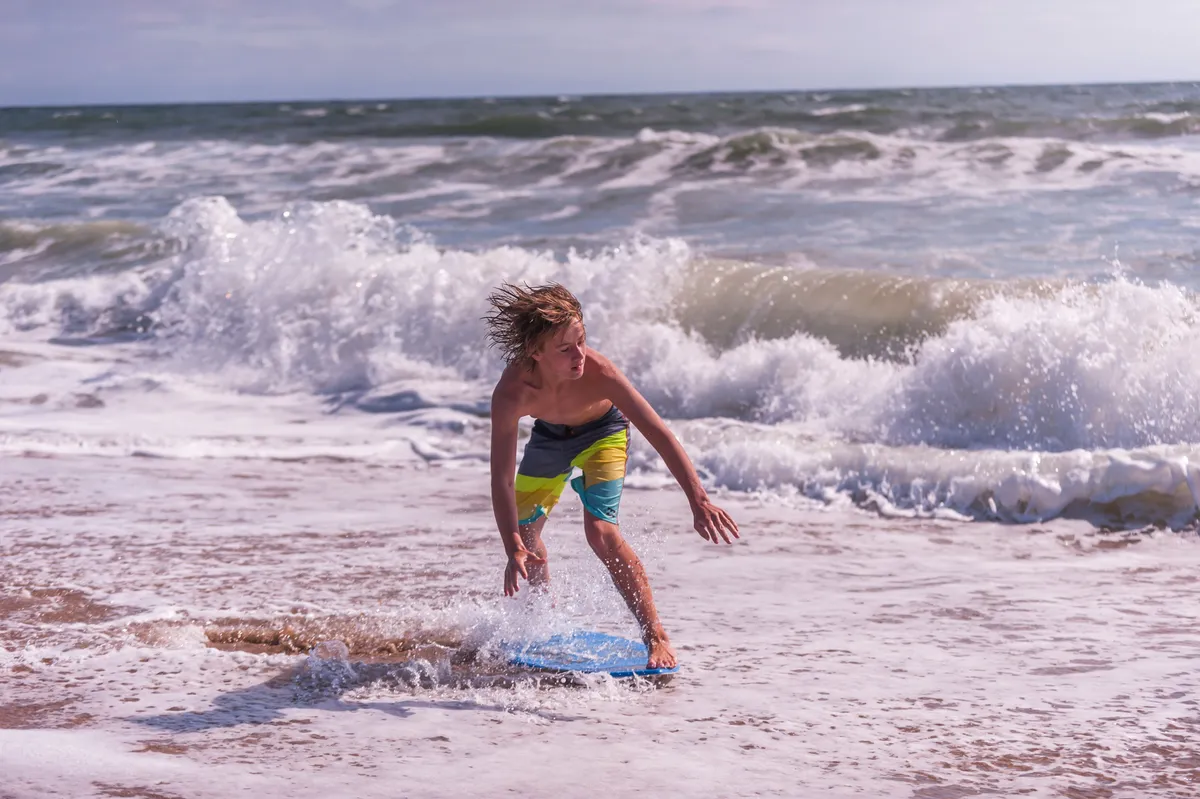 A playful beach scene featuring a young boy surfing through rolling waves with the sandy beach in the background. The mood is active and lively, capturing the spirit of adventure and enjoyment of water sports.