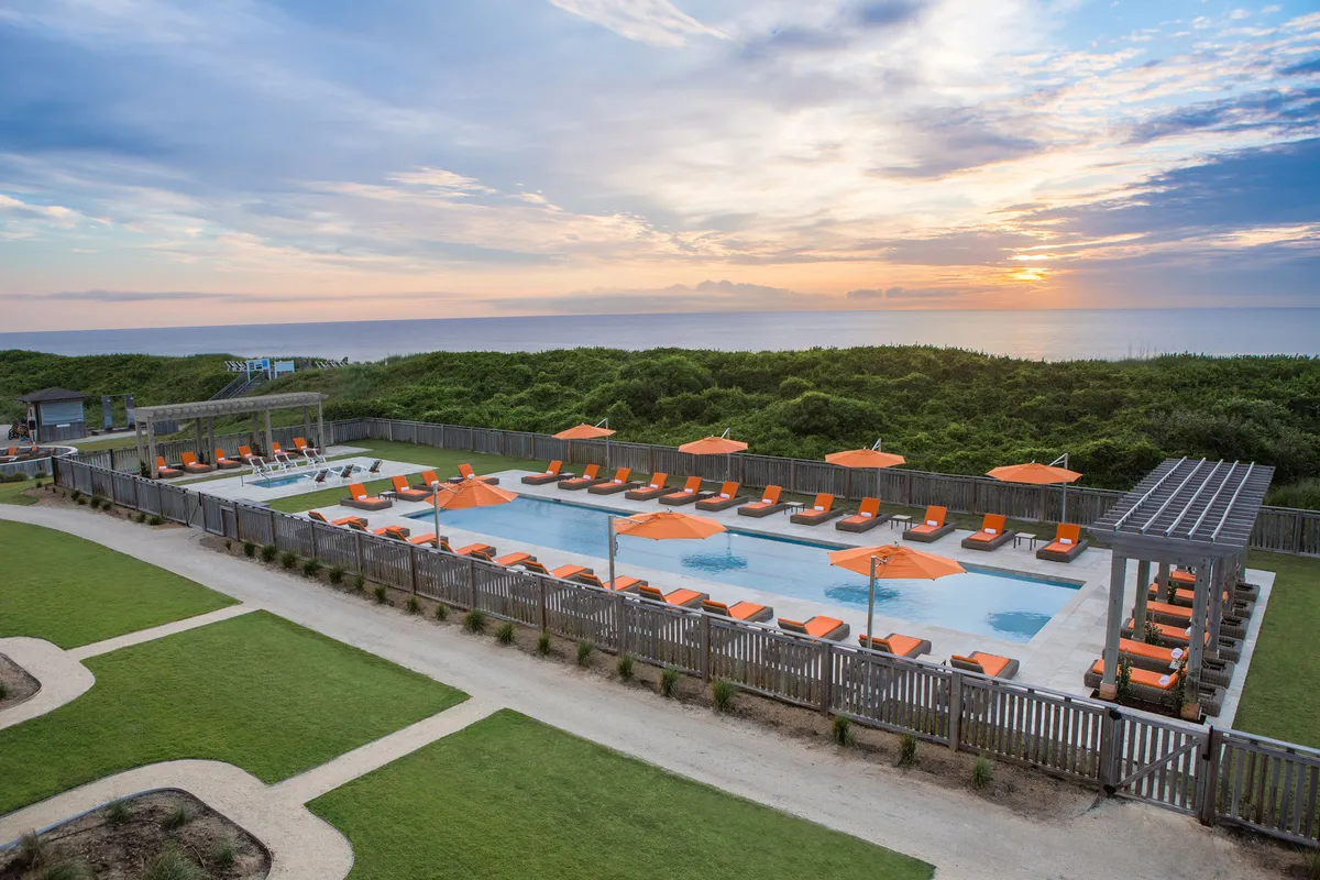 The image captures a tranquil pool scene at sunset, framed by comfortable lounge chairs and lush greenery. The water glistens softly, and the golden hues of the sunset create an uplifting, peaceful mood ideal for relaxation or social gatherings. This angle highlights the calm and inviting nature of the shared pool area during quiet evening hours.