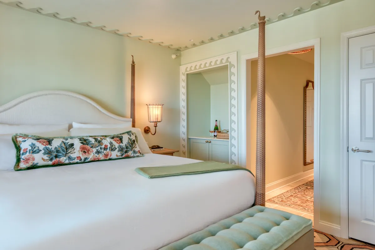 A soothing bedroom scene featuring a comfortable bed with a floral pillow set against a decorative wall, a sleek mirror, and a soft light fixture. The atmosphere is inviting and perfect for winding down after a day of activity.