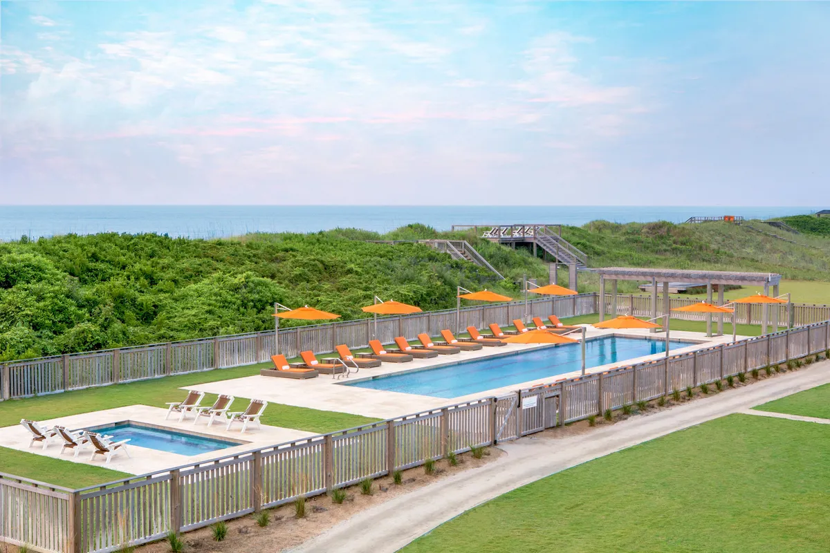A sunlit pool with inviting lounge chairs and vibrant orange umbrellas sits in the foreground, with an expansive ocean view beyond. The pool is surrounded by areas perfect for relaxation or casual gatherings, capturing the essence of laid-back luxury and seamless indoor-outdoor living.