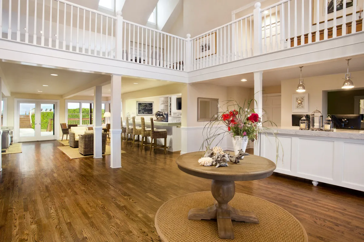 An airy interior with a large round table adorned with a bright floral arrangement, set in an open space with high ceilings and expansive windows. The view through the windows connects the indoors to the outdoor grounds, reinforcing themes of openness and tranquility. The environment is calm and inviting, suitable for gathering, contemplation, or wellness-focused activities.