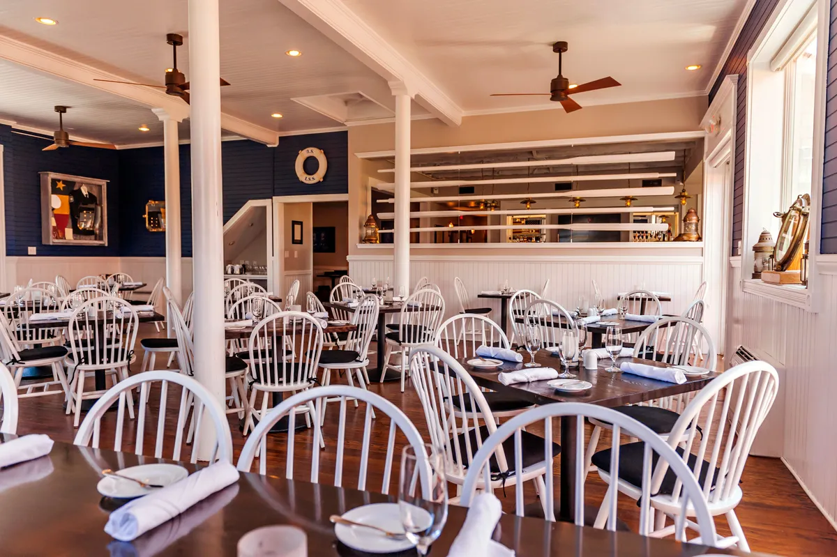 A welcoming indoor dining area featuring neatly arranged white chairs around several tables, a stylish bar area, and decorative wall art. The space radiates a relaxed, social atmosphere suitable for gathering with friends or family after a day of outdoor adventures.